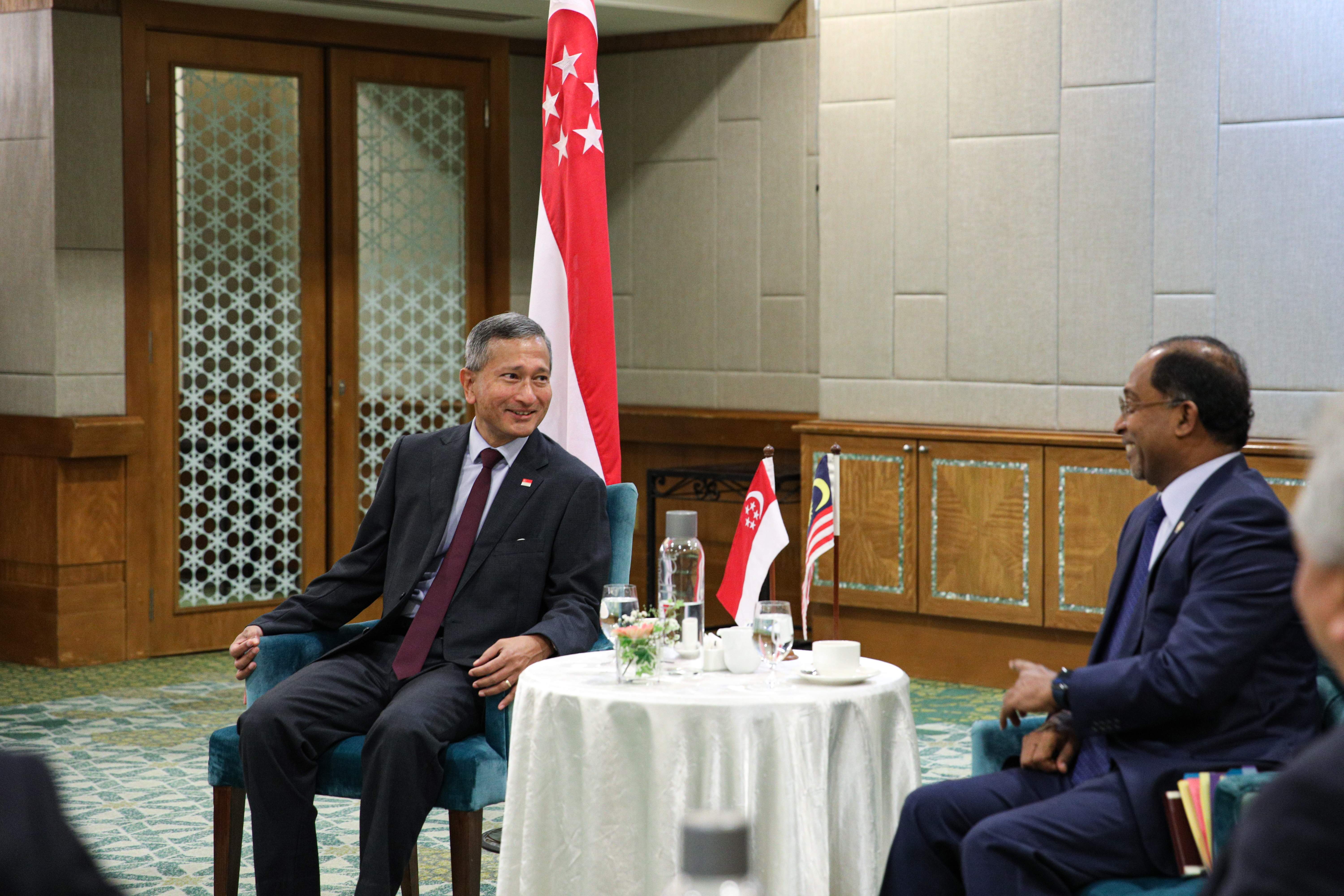 Two men in suits seated, facing each other. Singapore and Malaysia flags on table.